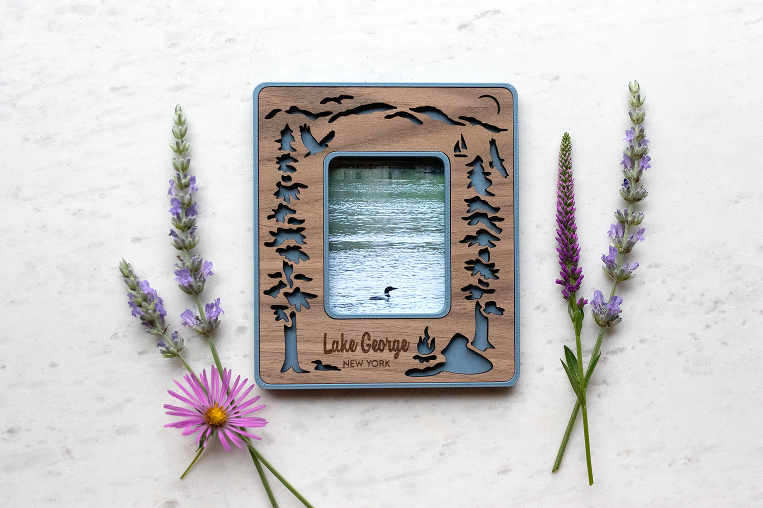 Lake George New York magnetic polaroid frame in Instax Mini frame size. The frame features laser cut Adirondack mountain, evergreen trees, loons, osprey, and kayakers on Lake George. The mini frame is lying next to wildflowers and displays a photo of a loon swimming on the lake.
