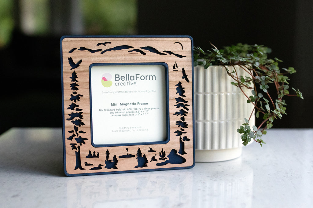 A standard Polaroid frame with dark blue plastic housing and laser cut cherry inlay. The frame has magnets on the back and is sitting next to a houseplant. The frame displays a card that reads "BellaForm Creative, Mini Magnetic Frame". The Polaroid frame fits standard 600 / SX-70 / iType photos.