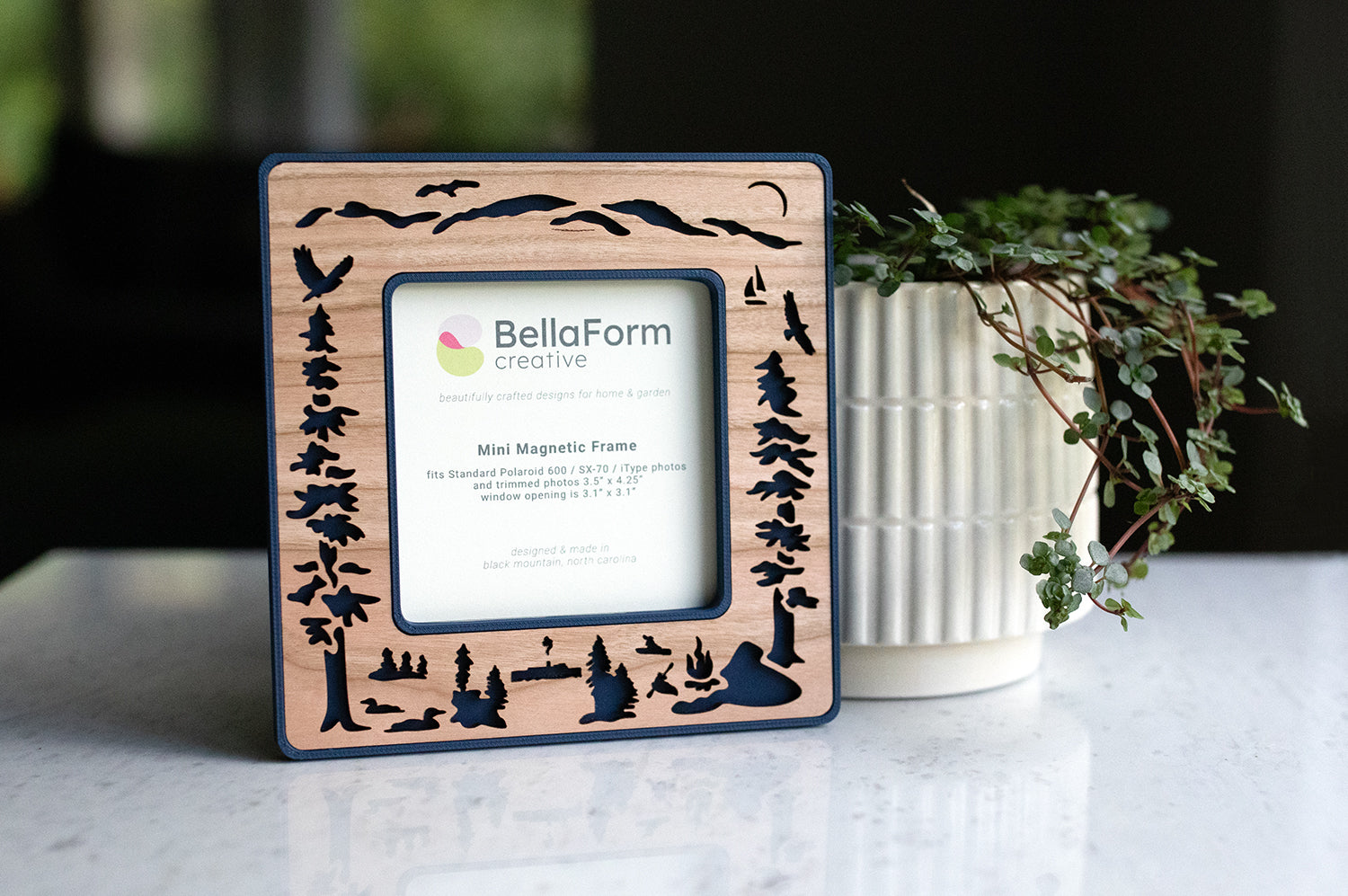 A standard Polaroid frame with dark blue plastic housing and laser cut cherry inlay. The frame has magnets on the back and is sitting next to a houseplant. The frame displays a card that reads "BellaForm Creative, Mini Magnetic Frame". The Polaroid frame fits standard 600 / SX-70 / iType photos.