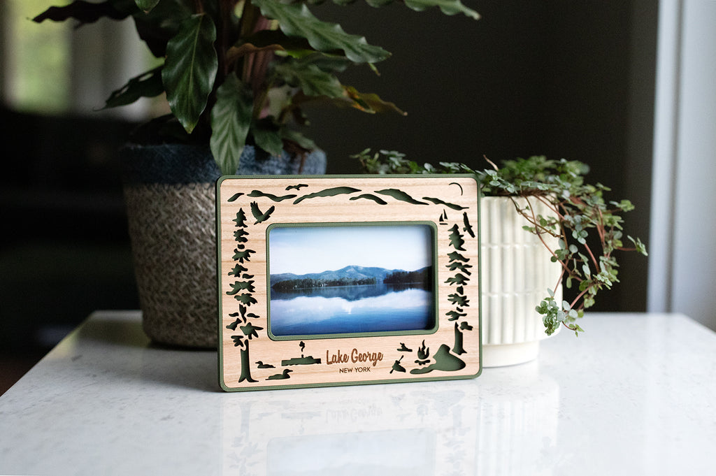 Instax Wide frame for Lake George lovers sitting upright next to a small houseplant. Frame design includes the Adirondack mountains, evergreen trees, and various wild bird species that are commonly found at the lake in upstate New York, including loons and osprey. A design for lake goers, lake houses, and lake lovers.