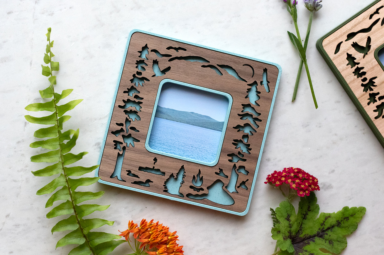 Polaroid Go frame in light blue with a laser cut walnut wood inlay, featuring Lake George New York loons, osprey, and mountains. The frame is surrounded by wildflowers and greenery and is lying on a white surface. Mini frame displays a trimmed photograph of Lake George and the Adirondack mountains in the distance.