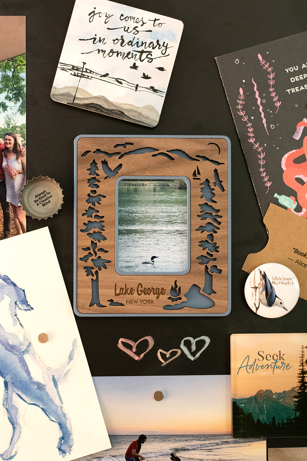 A magnetic Lake George Polaroid photo frame for Instax Mini hanging on a black, magnetic chalkboard. The frame has a walnut inlay and a slate blue border and includes the customized message: "Lake George New York". The frame is surrounded by postcards and photographs. All frames can be made with personalized messages.