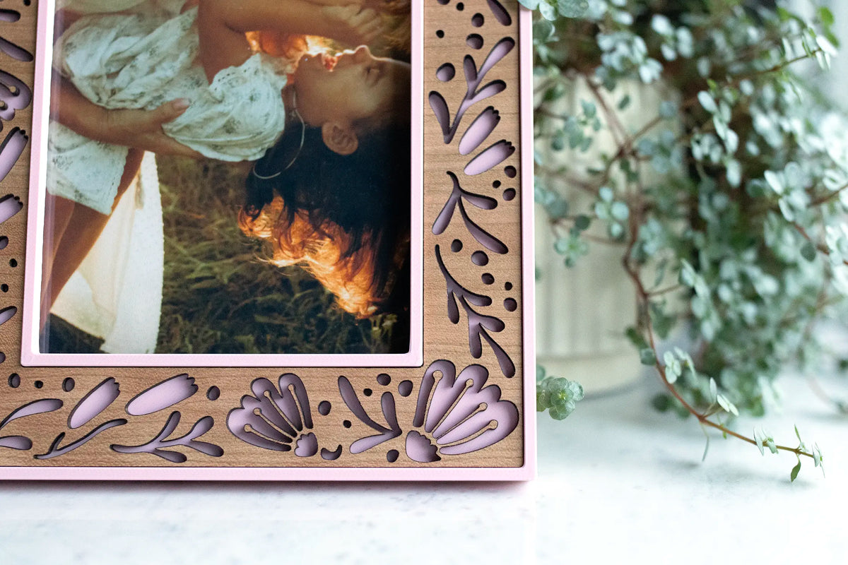 Close-up of the corner of a Camellia Pink picture frame showing laser-cut flower cutouts in a cherry wood inlay. The frame is holding a mother and daughter photo.