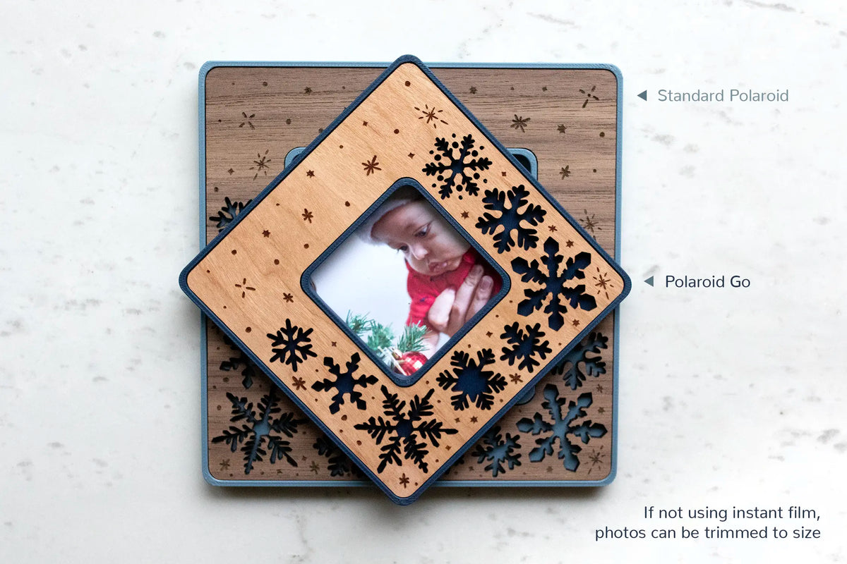 Two Polaroid-size snowflake magnetic frames stacked on white marble — a Standard Polaroid and the smaller Polaroid Go — with cherry and walnut wood inlays and laser-cut snowflakes, one holding a Christmas baby photo. Offered in walnut, cherry, or maple and cool winter colors, and personalizable for a stocking stuffer. Fits instant film, or trim a regular photo to size.