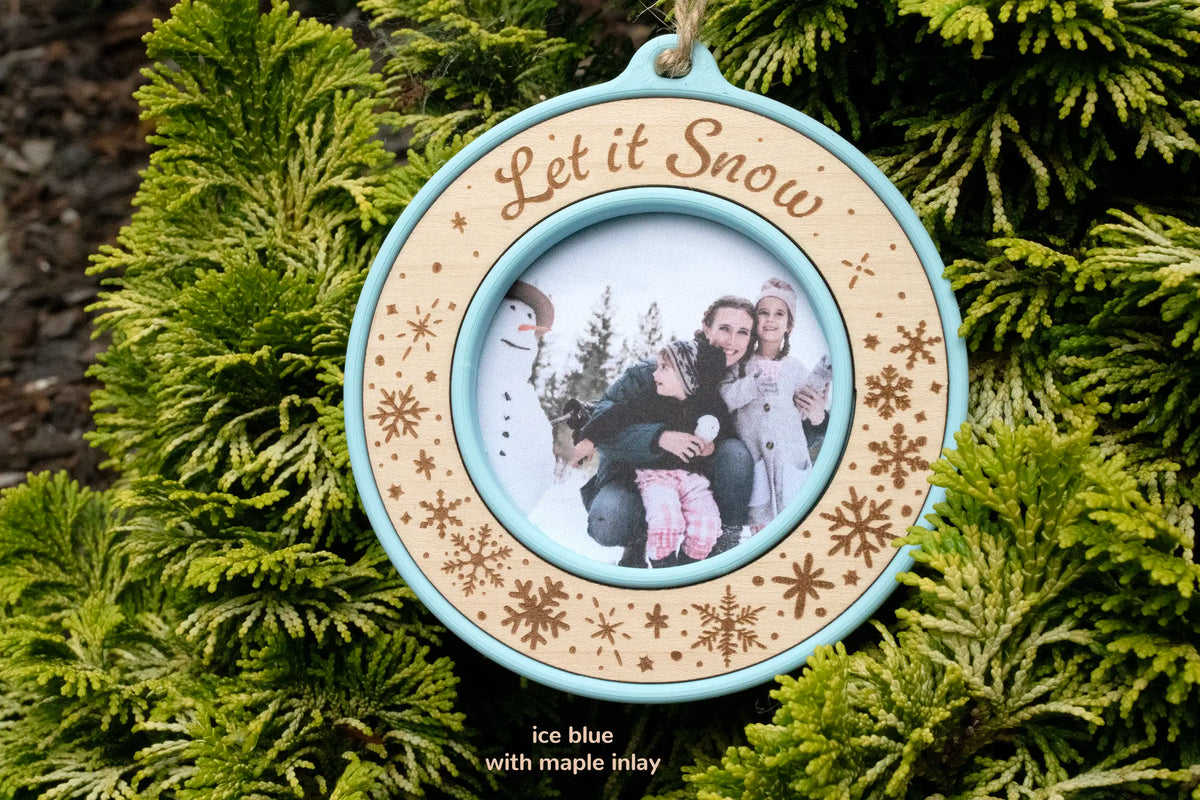 Snowflake photo ornament in ice blue with a maple inlay, laser-engraved "Let it Snow," holding a photo of a mother and two daughters beside a snowman, hanging on evergreen branches.