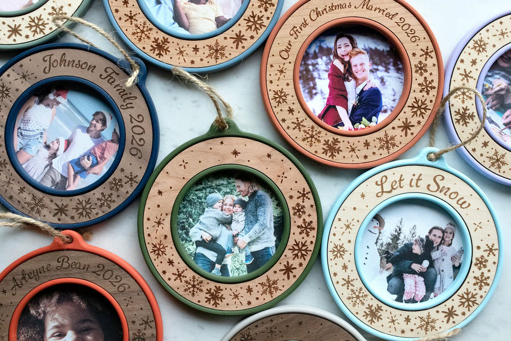 Overhead view of custom photo ornaments on white marble, centered on a text-free moss green and cherry ornament engraved only with snowflakes and holding a family photo. Around it are marine blue "Johnson Family 2026," terracotta "Our First Christmas Married 2026," ice blue "Let it Snow," and poppy red "Arleyne Bean 2026" mini picture frame ornaments.
