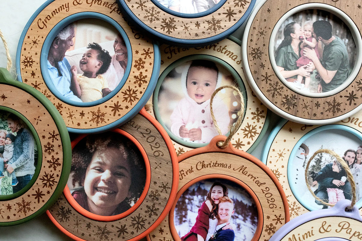Close-up of overlapping personalized Christmas photo ornaments in multiple colors, showing laser-engraved snowflakes on maple, cherry, and walnut inlays and round mini picture frames holding family, grandparent, baby's first Christmas, and newlywed photos.