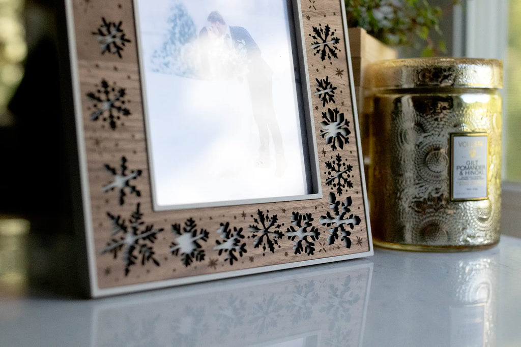 Detail of laser cut snowflake frame with walnut inlay and engraved floating snowflakes. The frame is linen white with walnut and is displayed next to a gold candle on a white marble counter.
