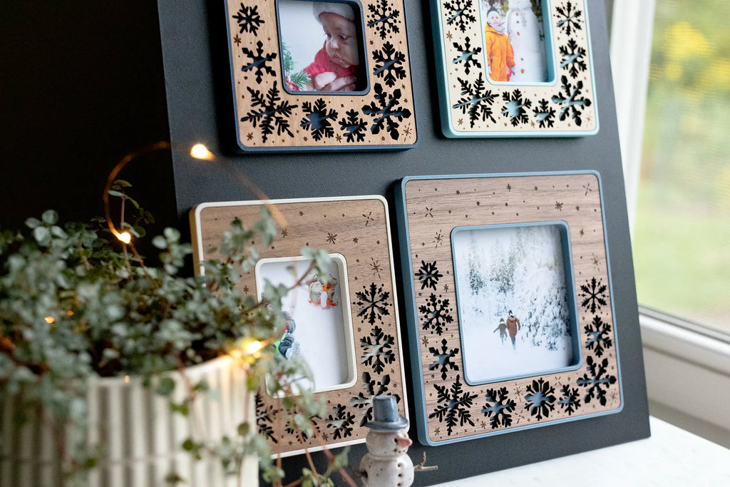Four Christmas snowflake magnetic frames on a black magnetic board, each holding a winter holiday photo, styled on a windowsill with a plant, fairy lights, and a ceramic snowman. The laser-cut wood inlays sit in a lightweight, durable 3D-printed housing, and each frame can be personalized with an engraved message or left snowflake-only.