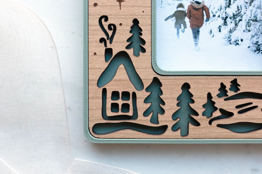 Close-up of a laser-cut cherry wood inlay showing a cabin with chimney smoke, pine trees, and snowy hills over a sage green 3D-printed housing.