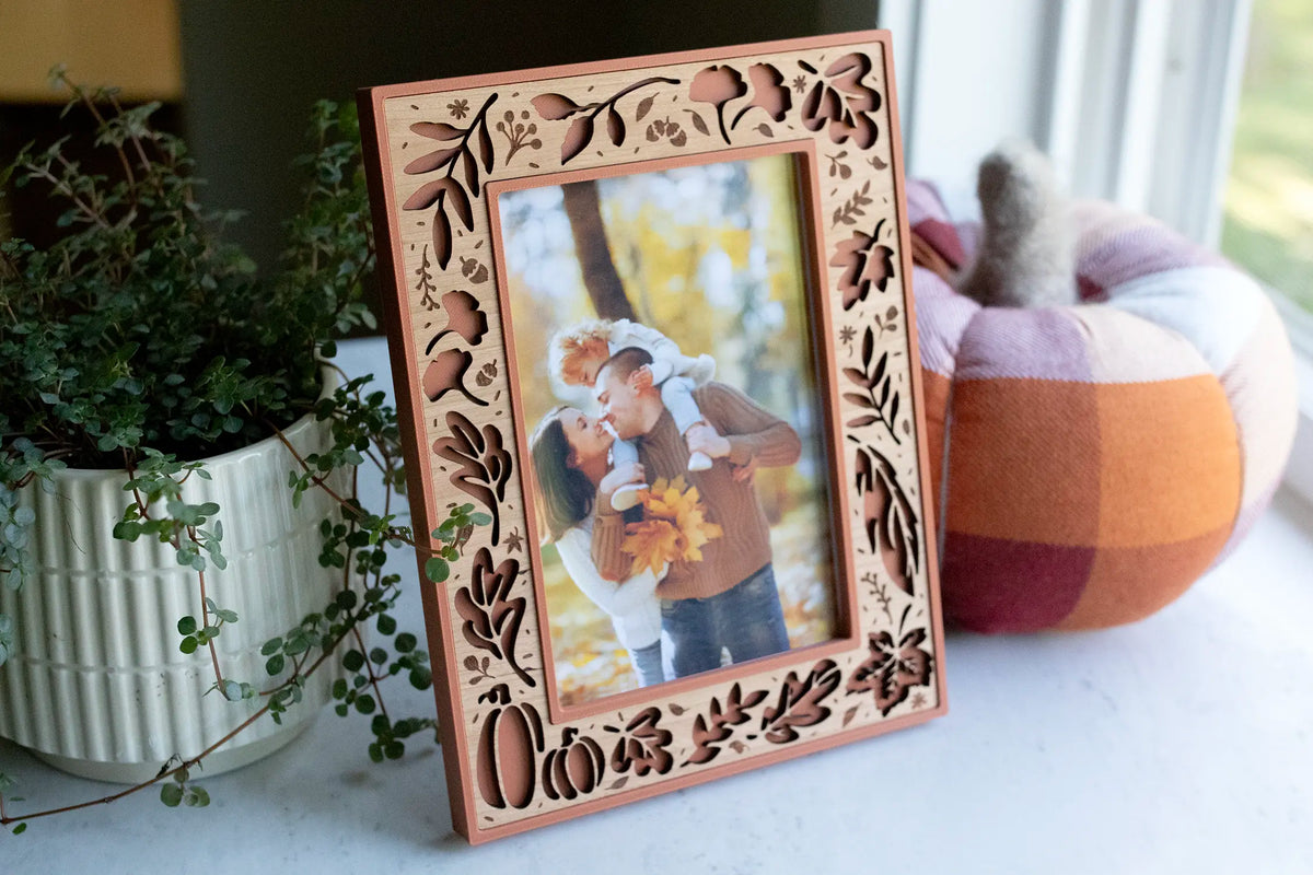 Personalized fall picture frame in terracotta with a cherry wood inlay, laser-cut with oak leaves, acorns, branches, and pumpkins — a custom autumn keepsake and Thanksgiving photo gift. Shown on a sunny windowsill holding a fall family portrait, styled with a plaid pumpkin and greenery.
