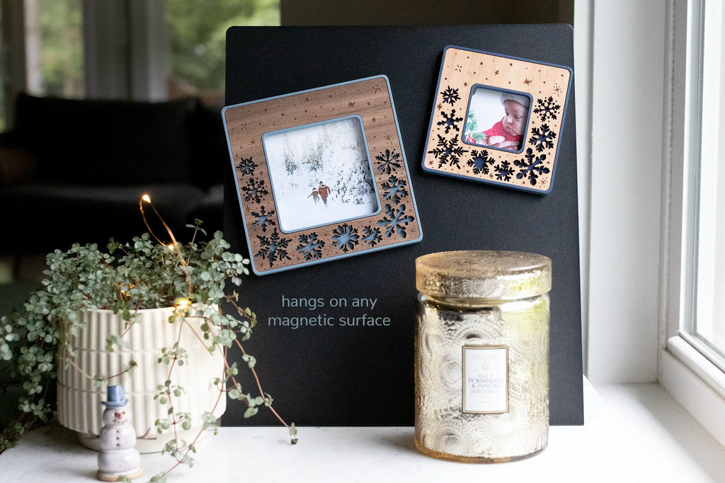 Two laser-cut snowflake magnetic frames on a black magnetic board, each holding a winter photo, styled on a windowsill with a plant, fairy lights, a candle, and a ceramic snowman. The walnut wood-inlay frames have blue cut-out snowflakes and can be personalized with an engraved message or left snowflake-only. More colors — shades of blue and white — and three woods (walnut, cherry, maple) available.