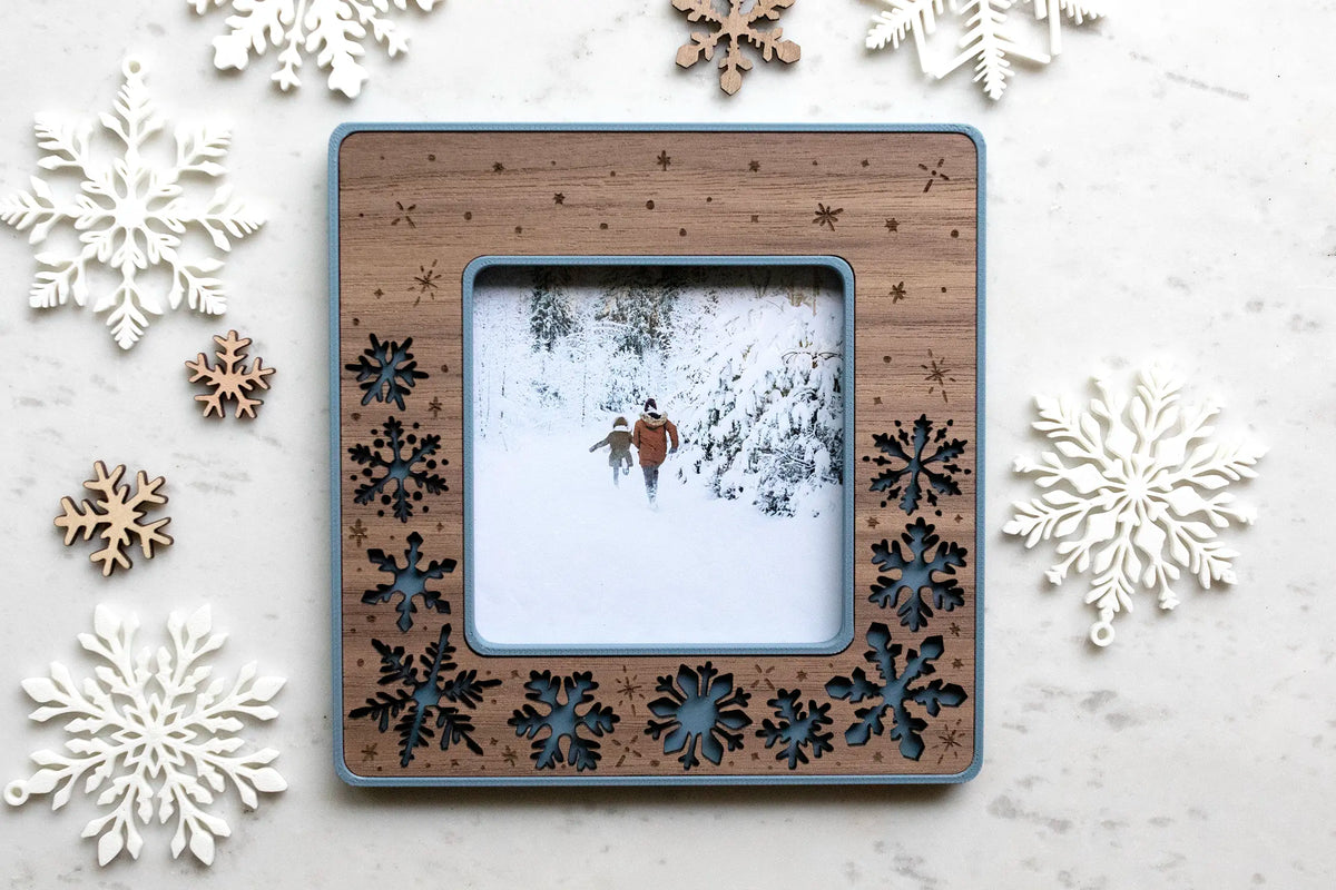 A Polaroid-size laser-cut walnut snowflake magnetic frame with blue frame housing, holding a winter photo of two people walking in the snow. Scattered wood and white snowflakes surround it on white marble. The frame is magnetic to stick on the refrigerator or any magnetic surface.