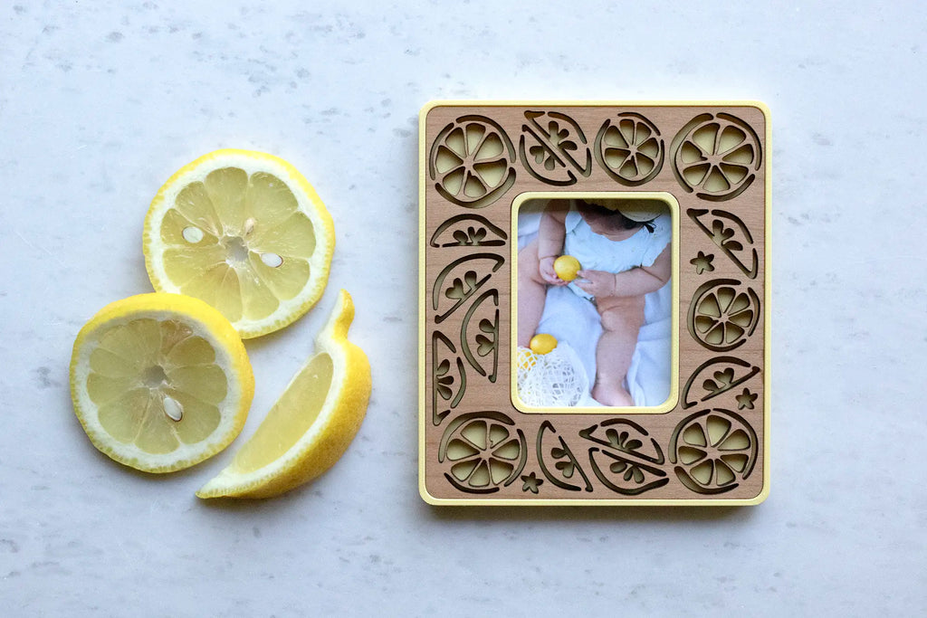 Magnetic, laser-cut lemon frame that holds an Instax Mini photo or a wallet size photo. The frame has a yellow housing with cherry wood laser-cut inlay. It is displaying a picture of a baby holding lemons and sitting next to lemon slices and wedges on a marble surface.