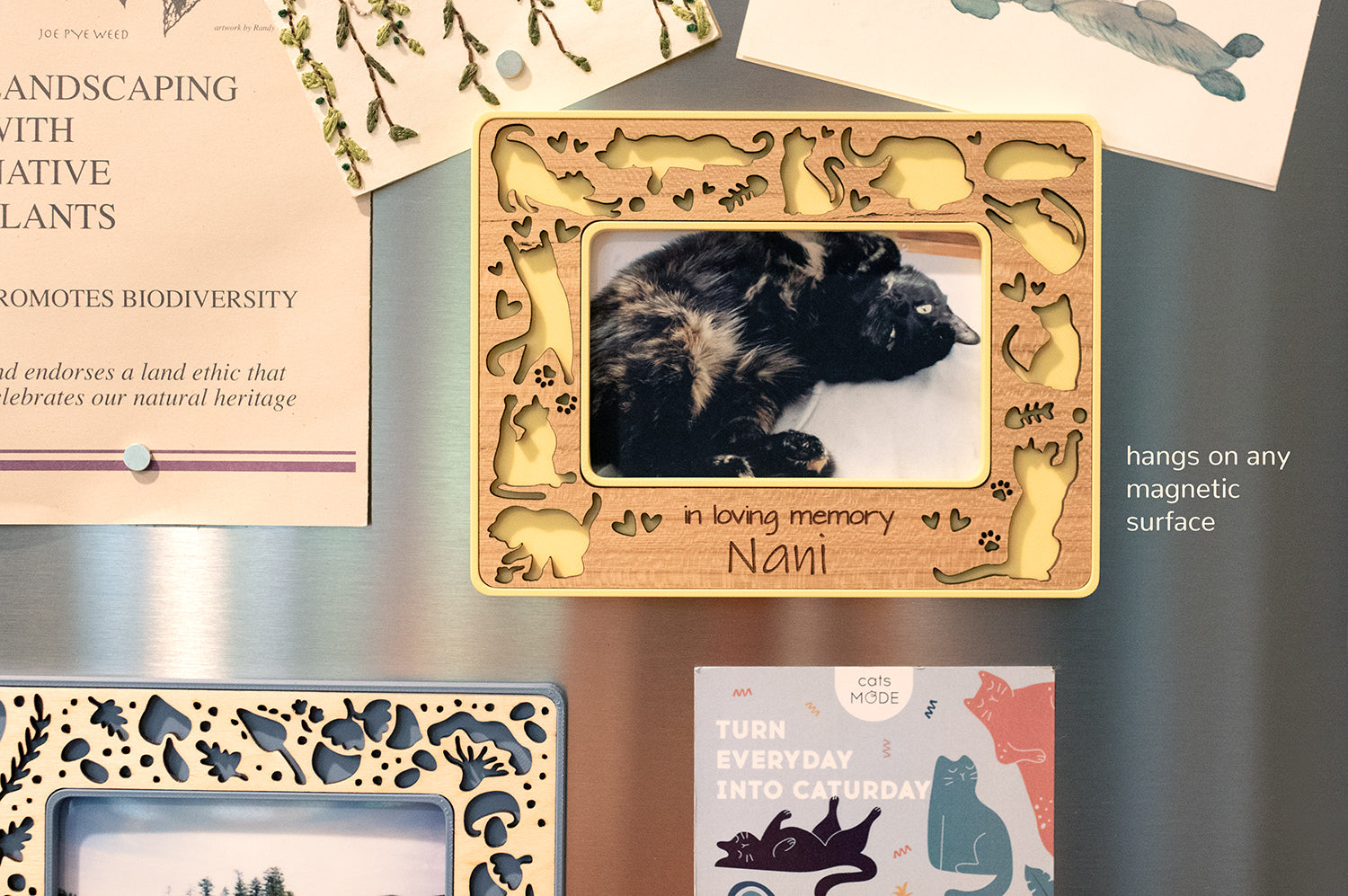 A yellow memorial cat frame hanging on a stainless steel refrigerator. The frame features cute laser cut cats doing various cute cat things, and there is an engraved message on the frame that reads "in loving memory Nani." A cat bereavement gift for cat lover.