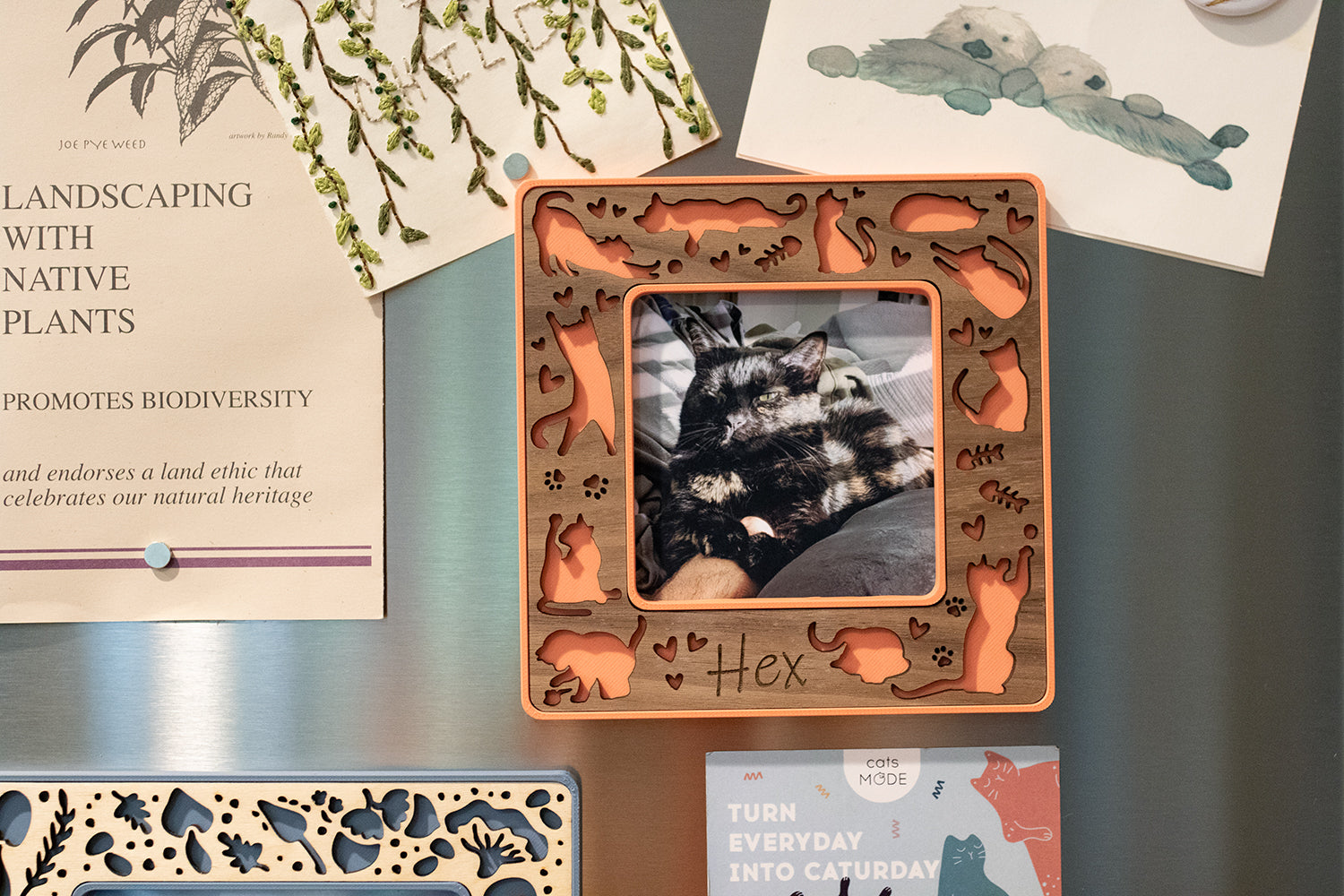 Magnetic frame for instant photo on refrigerator. Frame is coral pink with a walnut laser-cut cat inlay and displaying a photograph of a black cat.