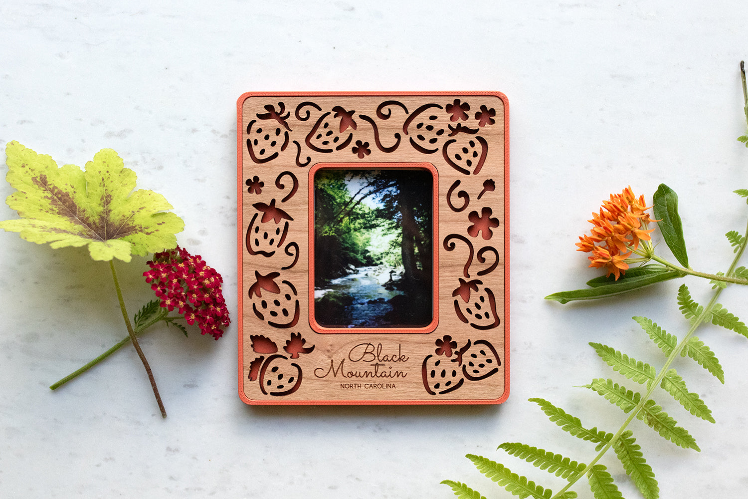 A magnetic strawberry polaroid frame keepsake made with a plastic housing and laser cut wooden inlay. The strawberry frame has a whimsical, laser cut design and a red border. The words "Black Mountain North Carolina" are engraved in the frame for personalization. Flowers are lying around the frame.