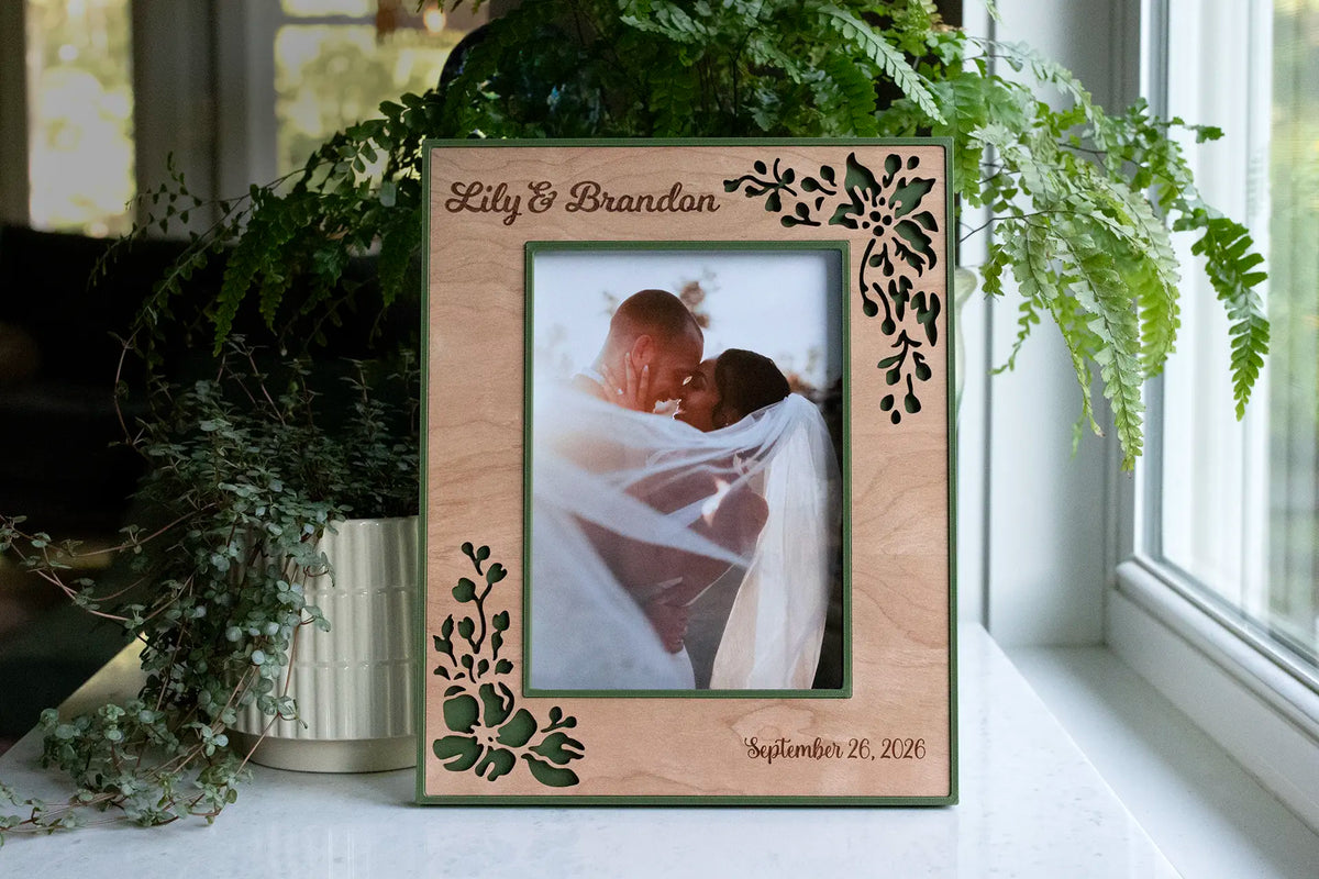 A personalized laser-engraved wedding picture frame featuring laser-cut flowers, a frame housing in the color of your choice, engraved names of the couple, and the date of their wedding or special event. Makes a thoughtful wedding gift or anniversary gift.