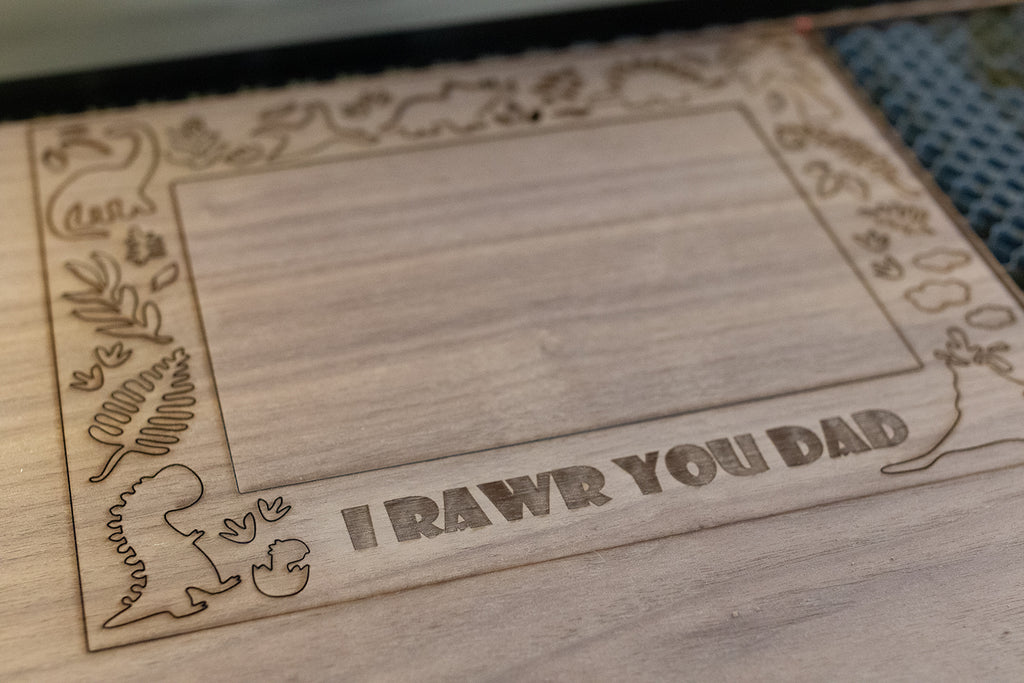 Wooden picture frame inlay with laser-cut dinosaur and plant designs lying on the laser cutting machine. The frame includes the engraving "I RAWR YOU DAD" in the wood.