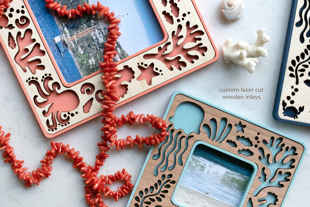 A close-up detail view of three mini magnetic sea creature frames. Frame designs include an octopus, crabs, starfish, jellyfish, sea turtles, fish, and ocean coral. The frames are pink coral, light blue, and navy with various wooden inlays. A coral necklace lies across the frames. There is a message that reads "custom laser cut wooden inlays".