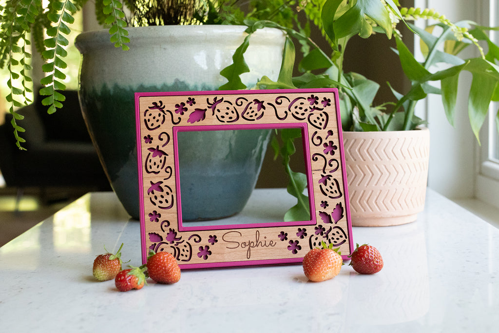 A detail view of the name "Maeva" laser engraved on a personalized picture frame. The frame features delicate strawberries and a bright pink frame with cherry wood inlay. It sits on a white surface with strawberries in the foreground.