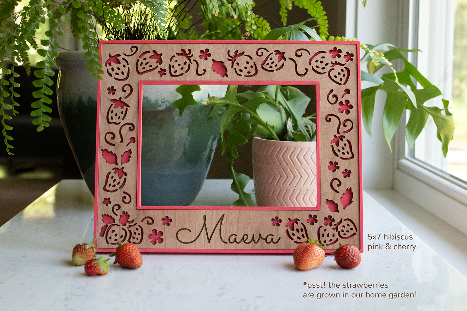 A whimsical strawberry frame featuring a bright pink frame and detailed laser cut strawberries in a custom cherry wooden inlay. The frame is sitting on a white counter and has greenery in the background.