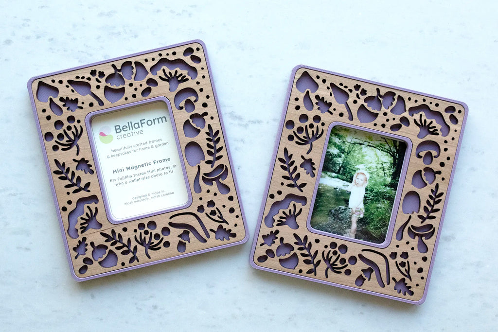 Two magnetic Instax Mini frames in lilac purple with cherry wood inlay and laser-cut mushroom, leaf, and fern cutouts, one with a BellaForm Creative insert card and one holding a photo of a child in a forest.