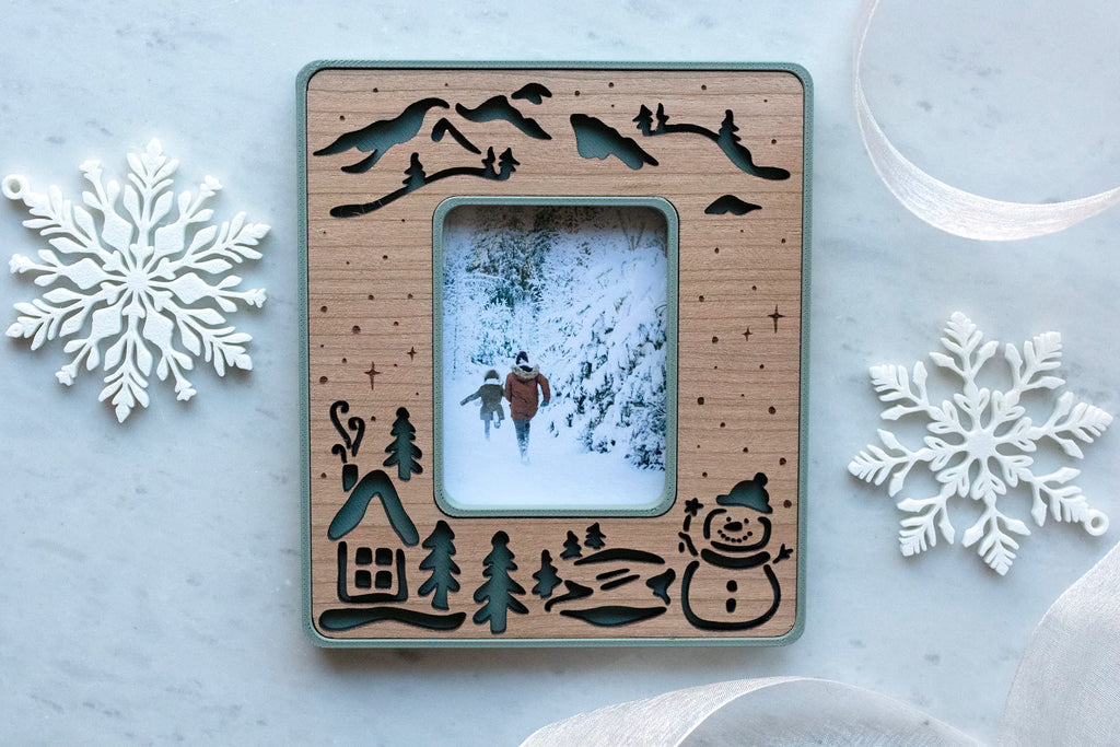 Sage green and cherry wood magnetic snowman picture frame with laser-cut snowman, cabin, evergreens, frozen lake, and snow-capped mountains under engraved falling snow, holding an Instax Mini photo of two people running through a snowy forest.