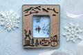 Sage green and cherry wood magnetic snowman picture frame with laser-cut snowman, cabin, evergreens, frozen lake, and snow-capped mountains under engraved falling snow, holding an Instax Mini photo of two people running through a snowy forest.
