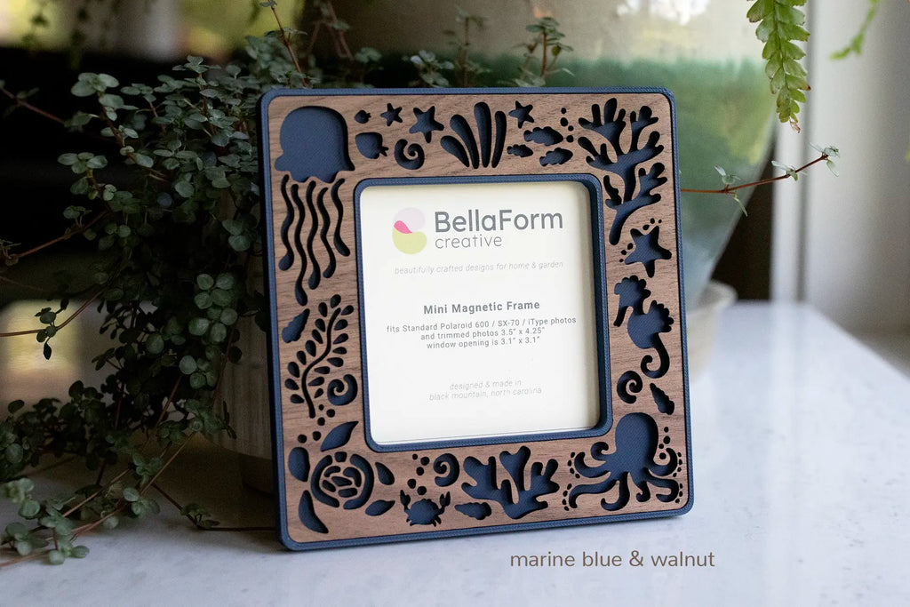 Hand crafted coastal frame with magnets for the refrigerator, sized for standard Polaroid photos. The frame features a laser-cut walnut inlay with octopus, starfish, jellyfish, sea turtle, seahorse, and coral. The frame is a dark marine blue color and is sitting next to greenery.