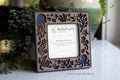 Hand crafted coastal frame with magnets for the refrigerator, sized for standard Polaroid photos. The frame features a laser-cut walnut inlay with octopus, starfish, jellyfish, sea turtle, seahorse, and coral. The frame is a dark marine blue color and is sitting next to greenery.