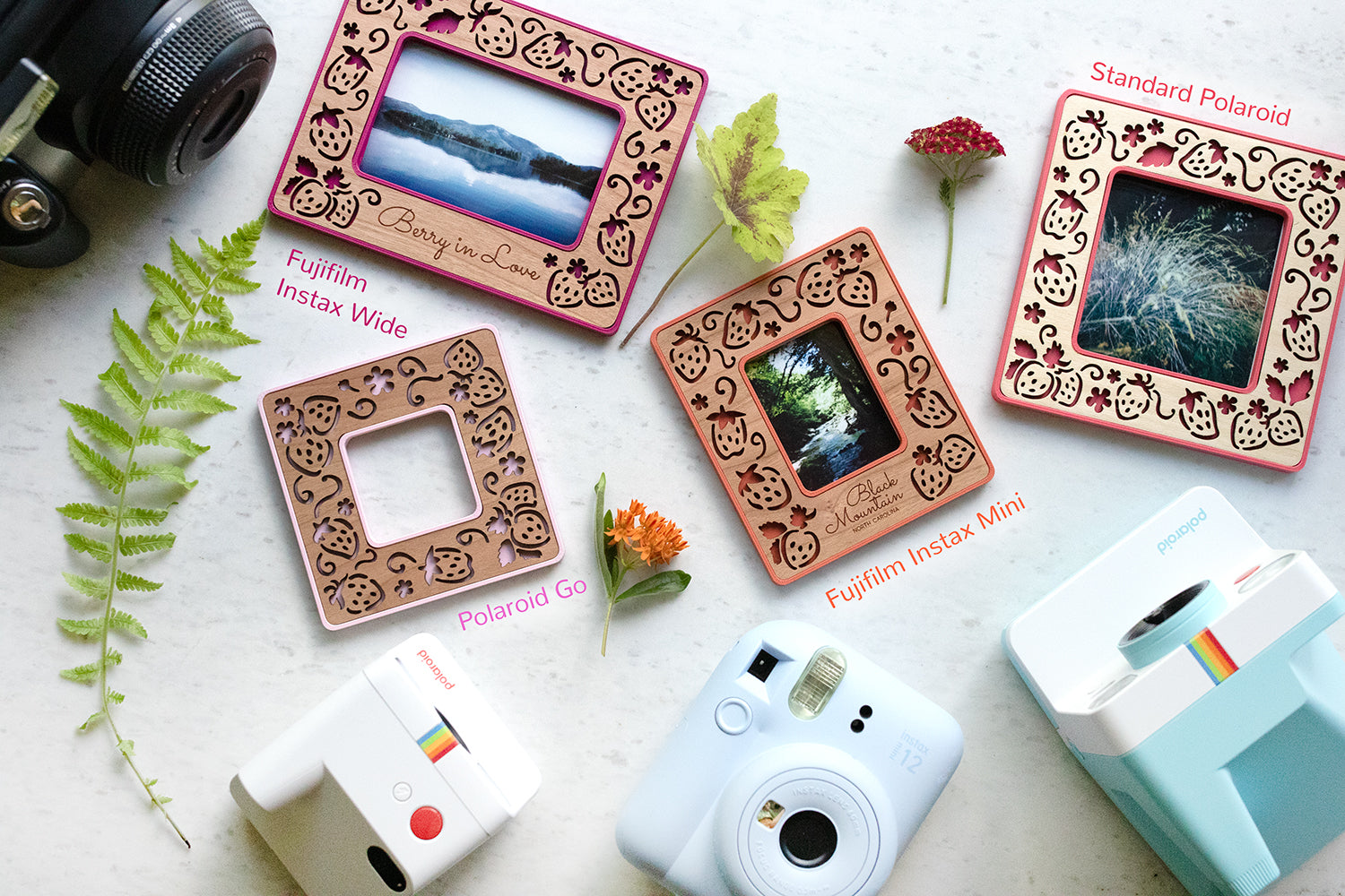 An assortment of colorful Polaroid photo frames including Instax Mini, Polaroid Go, Standard Polaroid, and Instax Wide. The frames are lying next to several different types of Polaroid and Fujifilm cameras that are used to take the photos. The strawberry frame design is shown in all four photo sizes in various shades of red and pink.