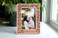Personalized cherry wood picture frame with a laser-cut mama and baby bear hugging amongst woodland flowers. The frame housing is camellia pink with engraved names "Kjalla & Mama" beneath the photo. The frame displays a photo of a mama holding her toddler daughter.