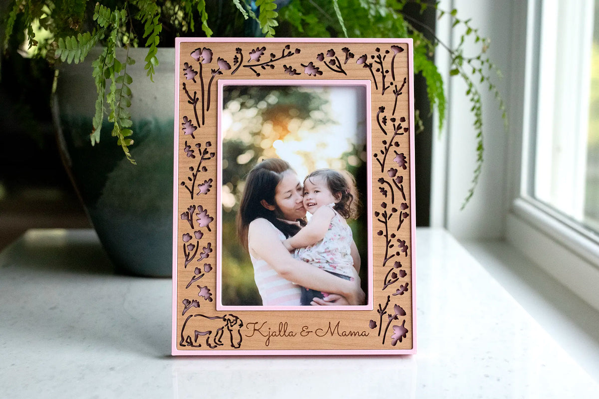 Personalized cherry wood picture frame with a laser-cut mama and baby bear hugging amongst woodland flowers. The frame housing is camellia pink with engraved names "Kjalla & Mama" beneath the photo. The frame displays a photo of a mama holding her toddler daughter.