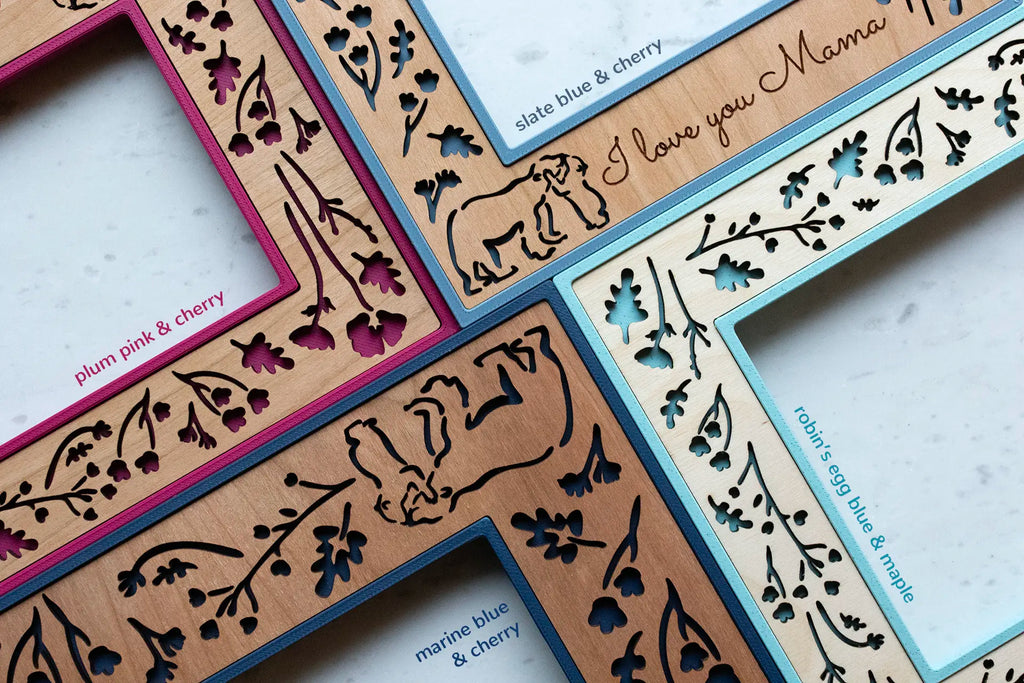 Close-up detail view of four colorful woodland laser cut frames with various wooden inlays showing wood grain and finish. Frames are various shades of blue and a bright plum pink. The laser cut design shows baby bear snuggling mama bear and surrounded by woodland flowers and leaves.