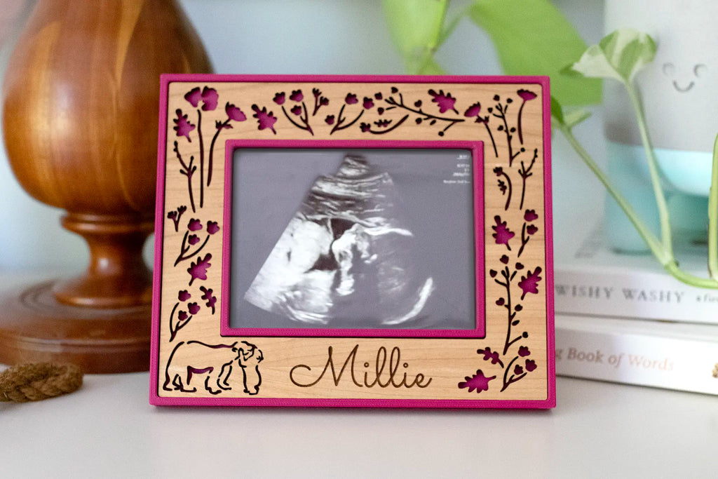 A personalized ultrasound picture frame holding a sonogram photo, with a laser-cut wildflower border and a mama bear and cub embracing at the bottom, engraved with the name "Millie," in plum pink and cherry wood. It stands on a shelf in a baby nursery.