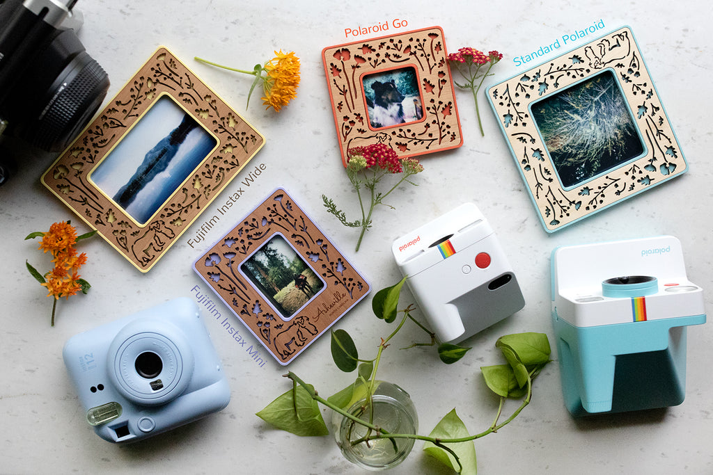 An assortment of colorful Polaroid photo frames including Instax Mini, Polaroid Go, Standard Polaroid, and Instax Wide. The frames are lying next to several different types of Polaroid and Fujifilm cameras that are used to take the photos.