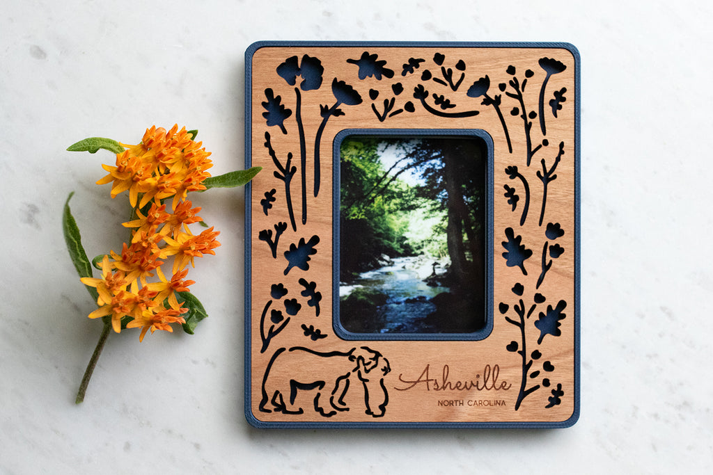 A magnetic polaroid photo frame with baby bear hugging mama bear. The design has a dark blue plastic housing with cherry wood inlay. The wooden inlay is laser cut with an intricate woodland flowers and bears design. The frame is personalized with the words "Asheville North Carolina". There is an orange flower lying next to the frame.