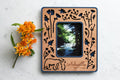 A magnetic polaroid photo frame with baby bear hugging mama bear. The design has a dark blue plastic housing with cherry wood inlay. The wooden inlay is laser cut with an intricate woodland flowers and bears design. The frame is personalized with the words "Asheville North Carolina". There is an orange flower lying next to the frame.