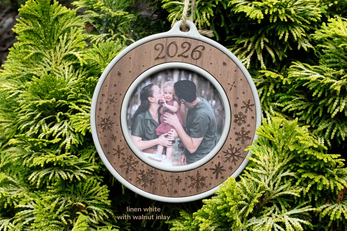 Keepsake Christmas photo ornament in linen white with a walnut inlay, laser-engraved "2026" with snowflakes, holding a photo of parents kissing their toddler's cheeks, hanging on evergreen branches.