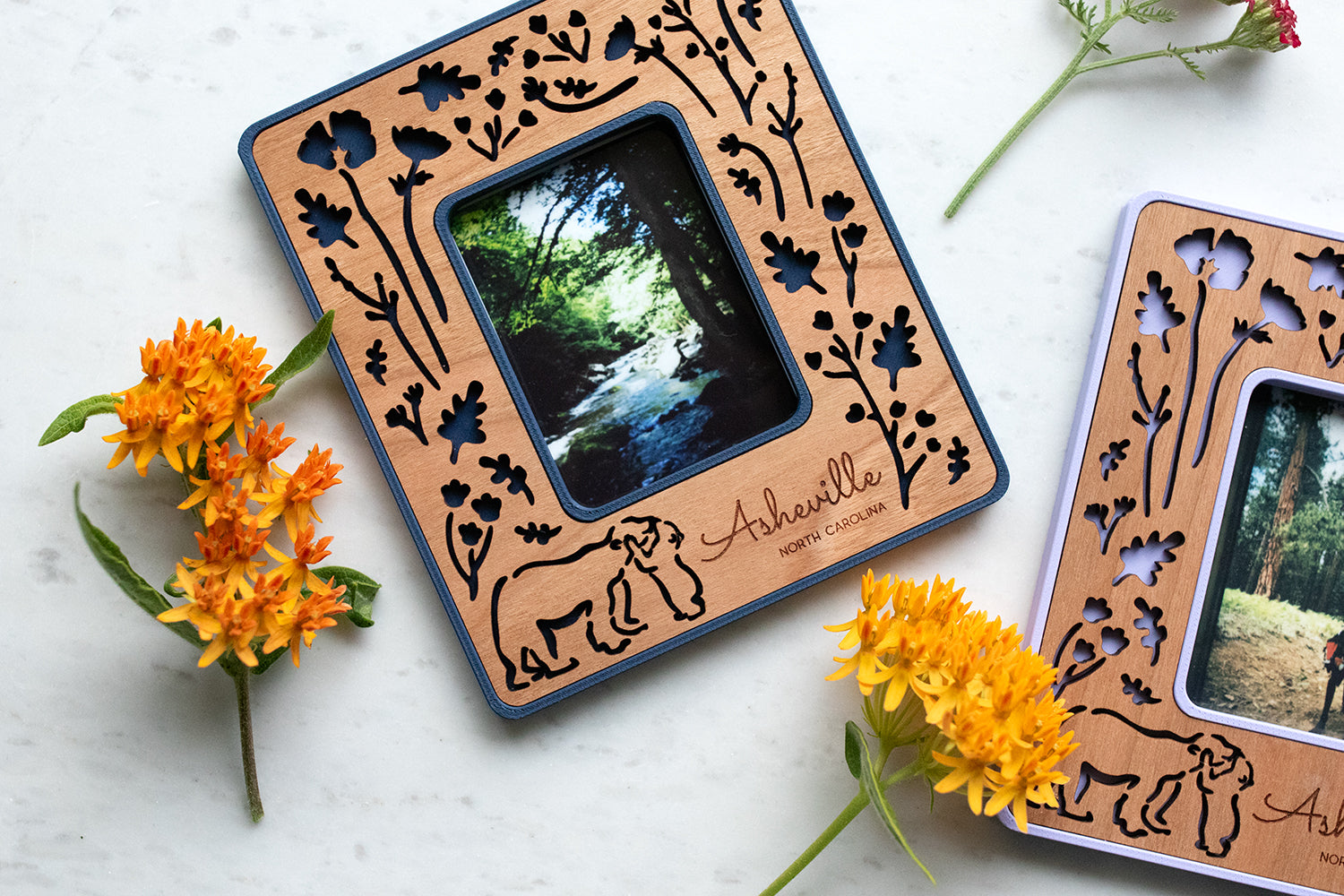 A mini photo frame that holds several common Polaroid and Fujifilm film sizes. The frames are magnetic and can be customized with engraved messages, names, and dates. Two frames in navy blue and periwinkle purple are lying on a white marble surface with flowers.