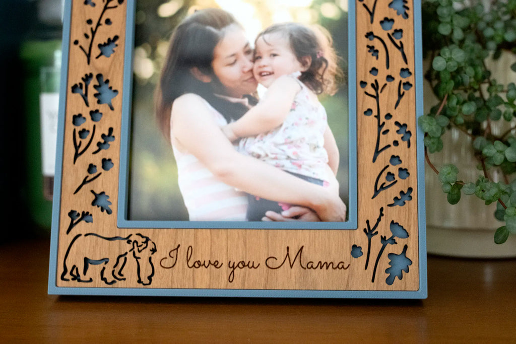 A close-up of a Mama and Baby Bear picture frame showing a laser-cut wood inlay of wildflowers over a slate blue housing, with a mama bear and cub outline beside the engraved words “I love you Mama.”