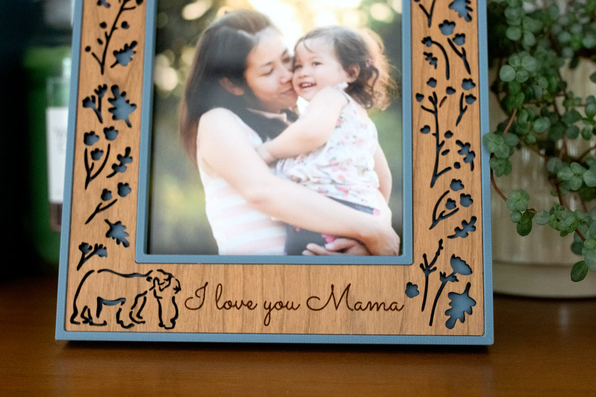 A close-up of a Mama and Baby Bear picture frame showing a laser-cut wood inlay of wildflowers over a slate blue housing, with a mama bear and cub outline beside the engraved words “I love you Mama.”