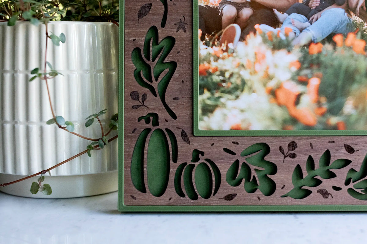 Detail view of a handmade fall picture frame in deep moss green with a laser-cut walnut wood inlay, showing pumpkins, oak and maple leaves, branches, and berries around the photo opening. A natural autumn and Thanksgiving decor piece, styled beside a white ribbed planter with trailing greenery.