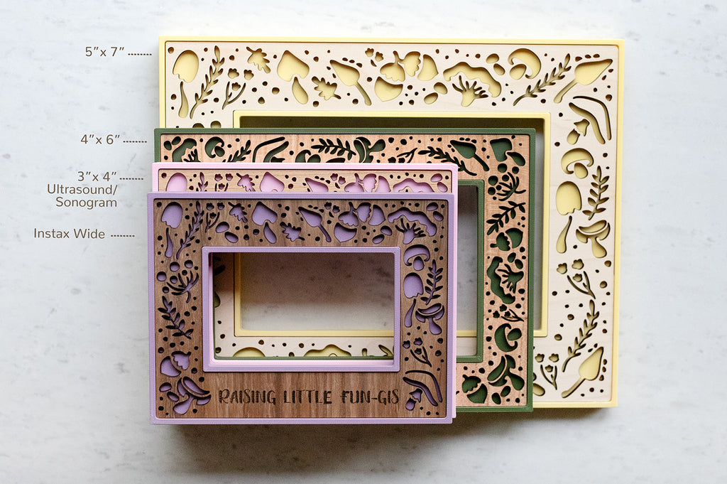 Four custom laser cut mushroom picture frames in a stack. Frames are green, yellow, purple, and pink with various wood types. The frames feature intricate laser cut designs with mushrooms and acorns. Image shows various frame sizes, including dimensions. 5x7 picture frame, 4x6 picture frame, 3x4 ultrasound picture frame, and Fujifilm Instax Wide picture frame.