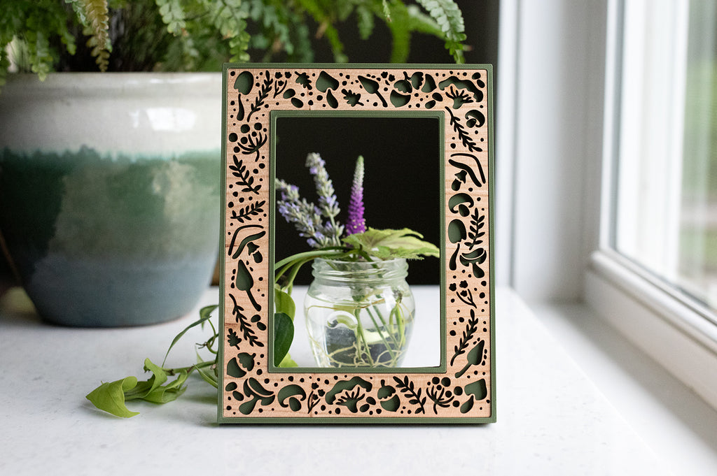 A green laser cut mushroom picture frame with intricate cherry wood inlay. The frame has a whimsical aesthetic and is surrounded by greenery and flowers. 4x6 picture frame with jar of flowers visible through the frame.