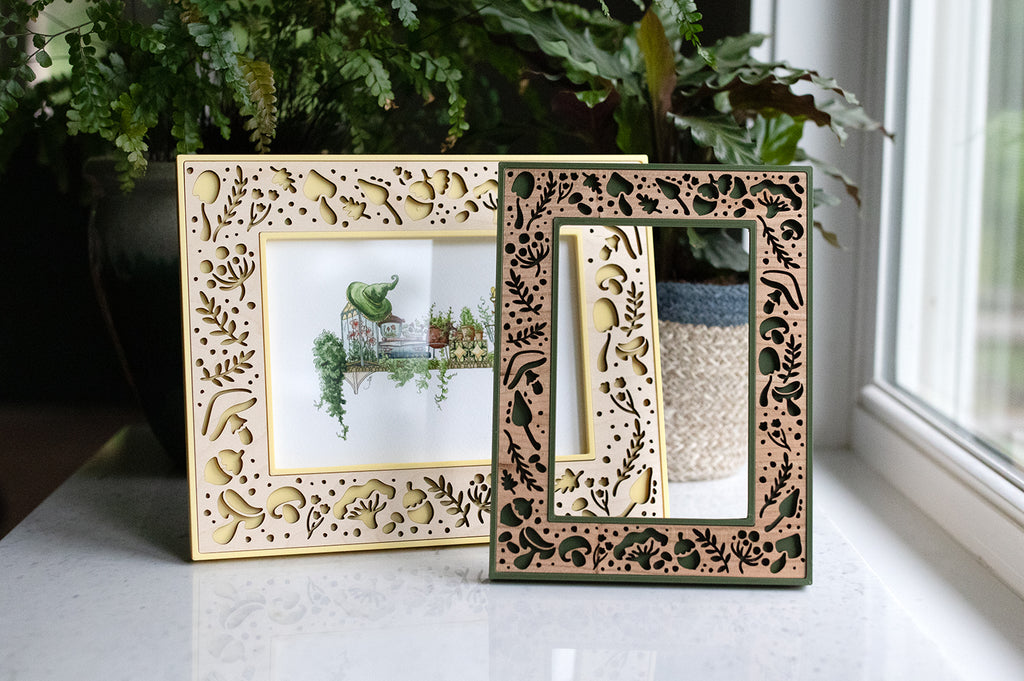 Whimsical mushroom picture frames. Included are a yellow and maple 5x7 picture frame and a moss green and cherry 4x6 picture frame. The yellow and maple frame display a painting of greenery for a garden witch's shelf. There is greenery in the background.