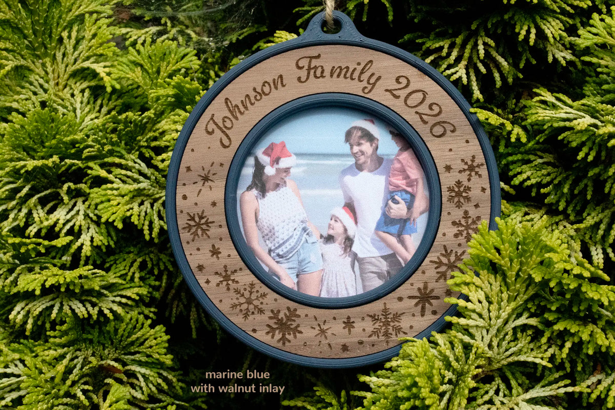 Personalized family Christmas ornament in marine blue with a walnut inlay, laser-engraved "Johnson Family 2026" with snowflakes, a mini picture frame holding a beach photo of a family of four in Santa hats, hanging on a Christmas tree.