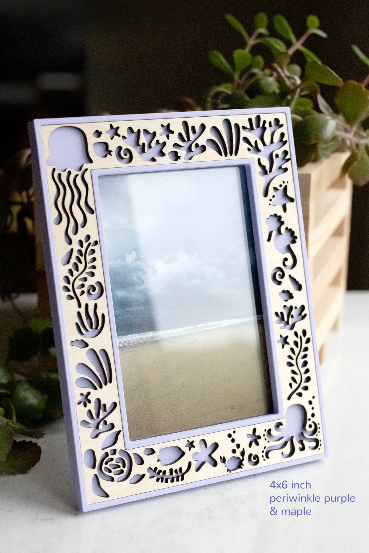 A purple and maple wood custom picture frame featuring laser cut sea creatures for an Under the Sea decor theme. Size is a 4x6 frame in vertical orientation, and it is displaying a photograph of the ocean. The frame is sitting on a white marble surface with greenery in the background.