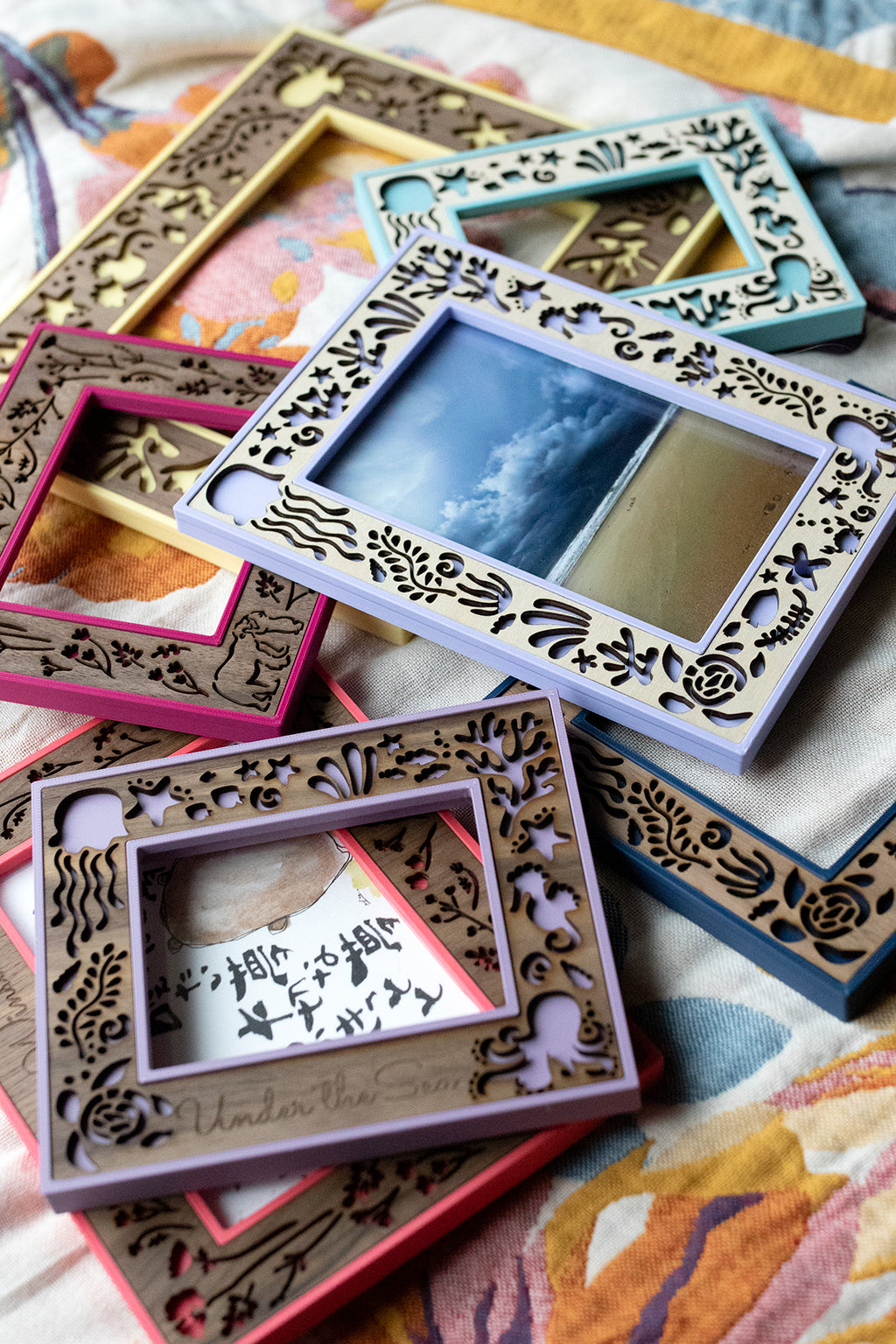 An assortment of colorful 3D printed and laser cut wooden ocean creatures themed picture frames laying on a colorful blanket. The intricately detailed laser cut designs incorporate several popular ocean creatures. Frames can be made in walnut, cherry, or maple wood.